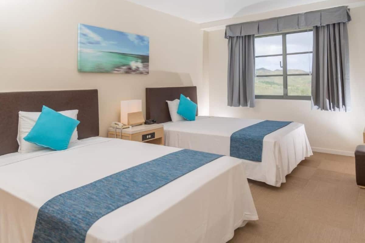Habitación de hotel con dos camas, una mesita de noche, una lámpara y una ventana con cortinas.