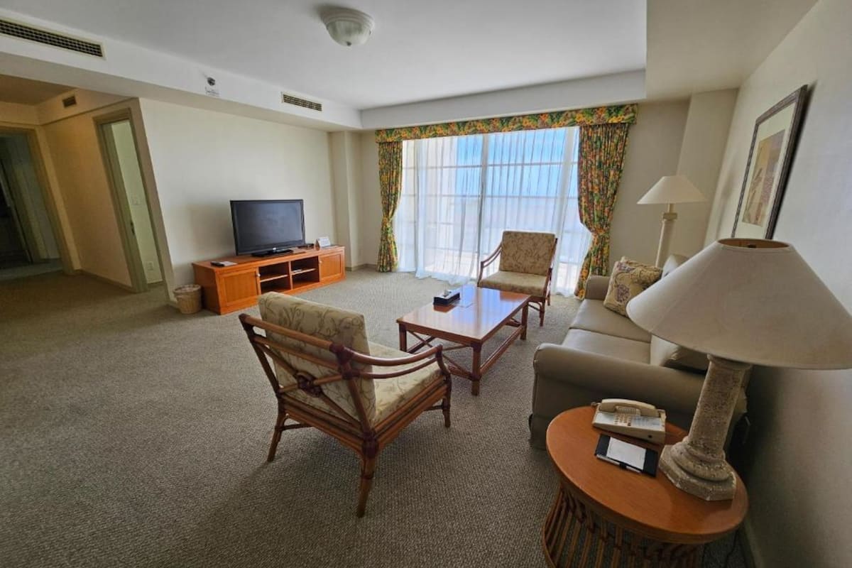 Habitación de hotel con televisión, dos sillones, un sofá, una lámpara y una mesita.
