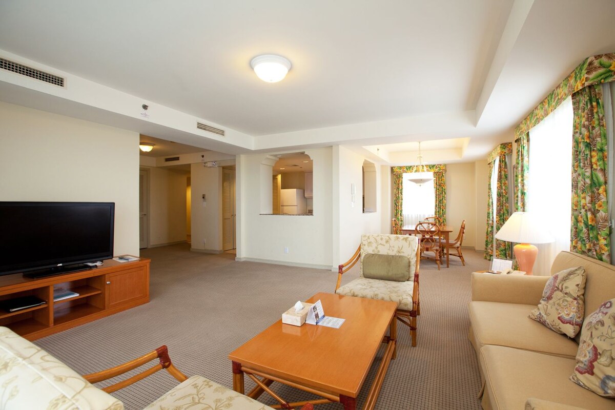 A hotel room with a flat-screen TV, a wooden coffee table, a sofa, a dining area with a table and chairs, and a window with floral curtains.