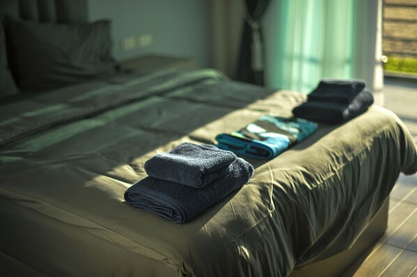 A neatly made bed with folded towels and a blanket, a window with curtains, and a view of greenery outside. - Reboot Retreat (Tha Sala)