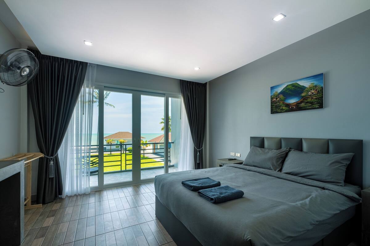 A modern hotel room with a large bed, a desk, and a view of the sea through a sliding glass door.