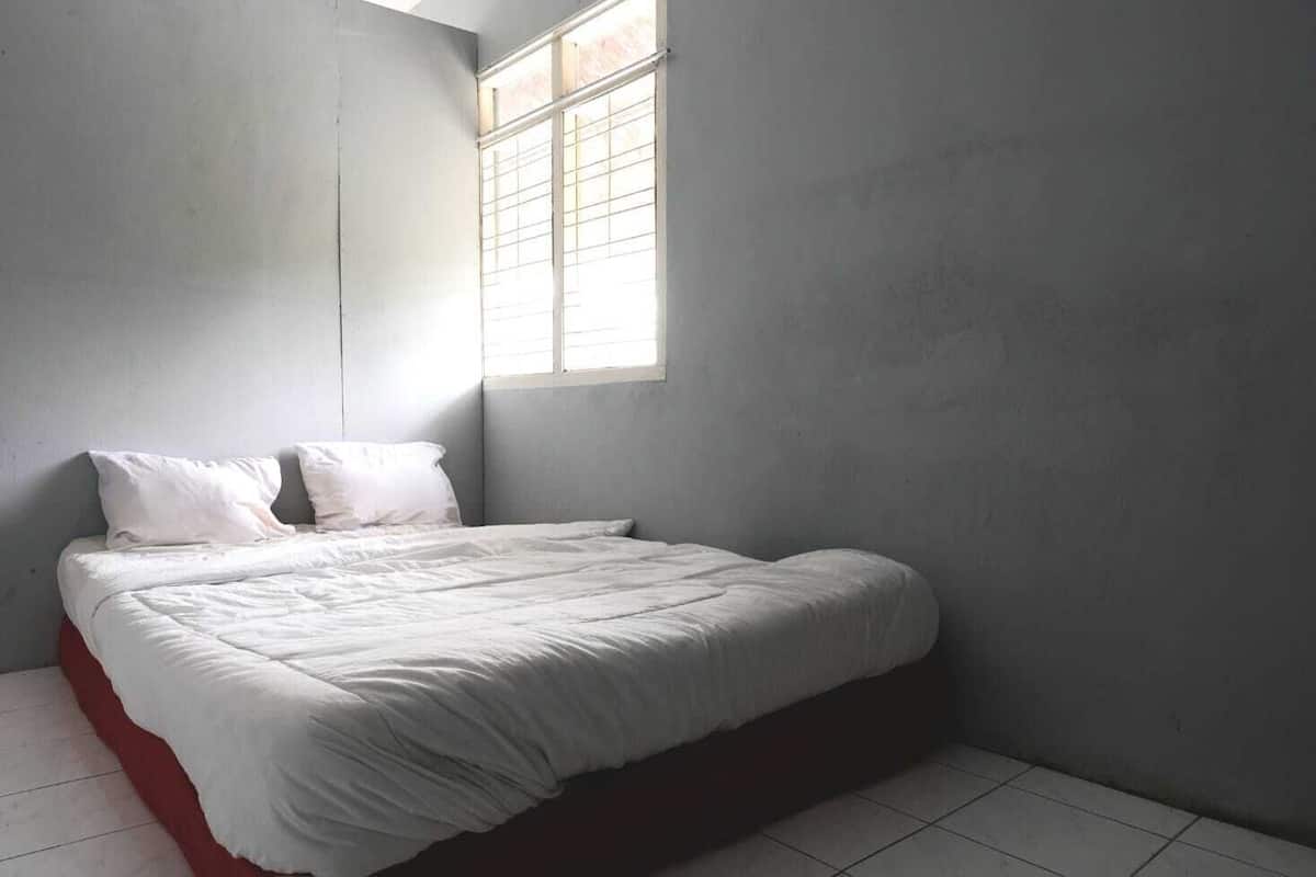 Een slaapkamer met een bed, witte beddengoed, een raam met zonwering en een effen muur.