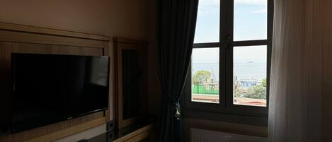 A hotel room with a bed, a TV mounted on a wooden cabinet, and a window with a view of the sea and buildings.