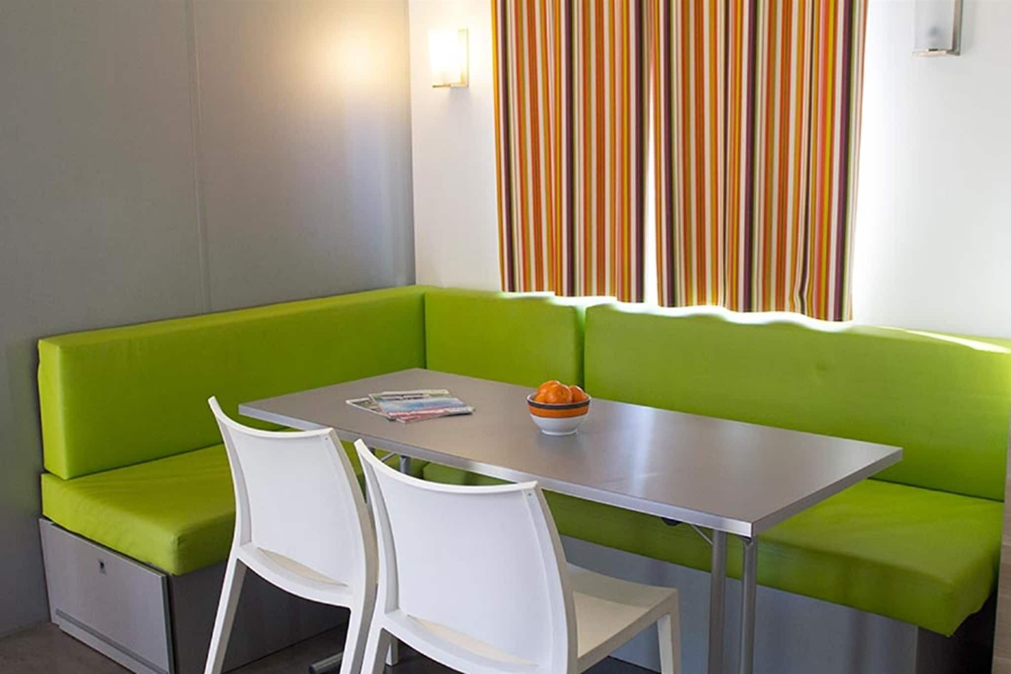 Una pequeña kitchenette con un sofá verde, sillas blancas y una mesa con un cuenco de fruta.