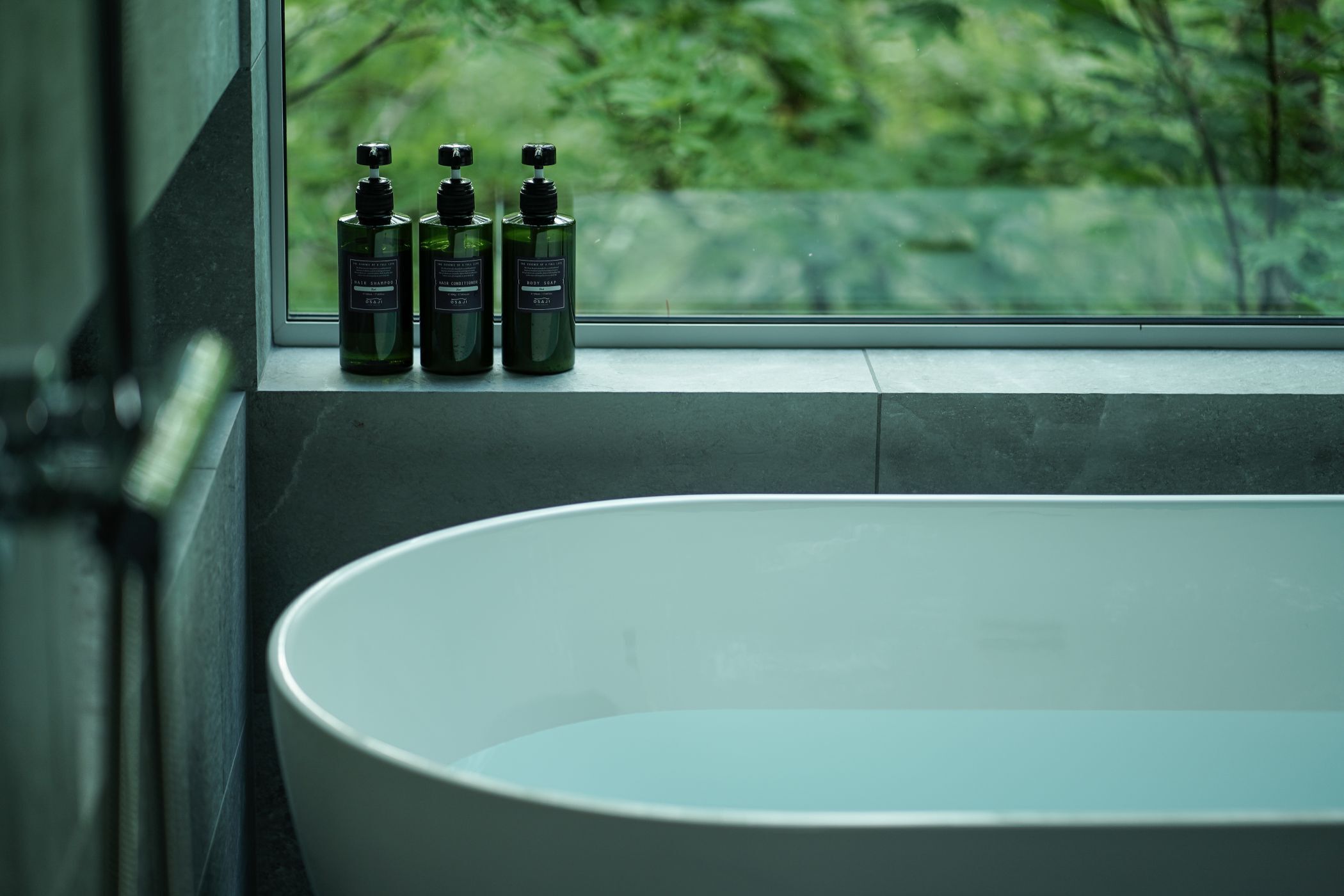 A bathroom with a large bathtub, a window with a view of greenery, and three bottles of shampoo on the windowsill.