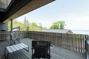 Une terrasse en bois avec une chaise Coleman et une table, donnant sur une zone boisée et un bâtiment doté d’un toit en pente.