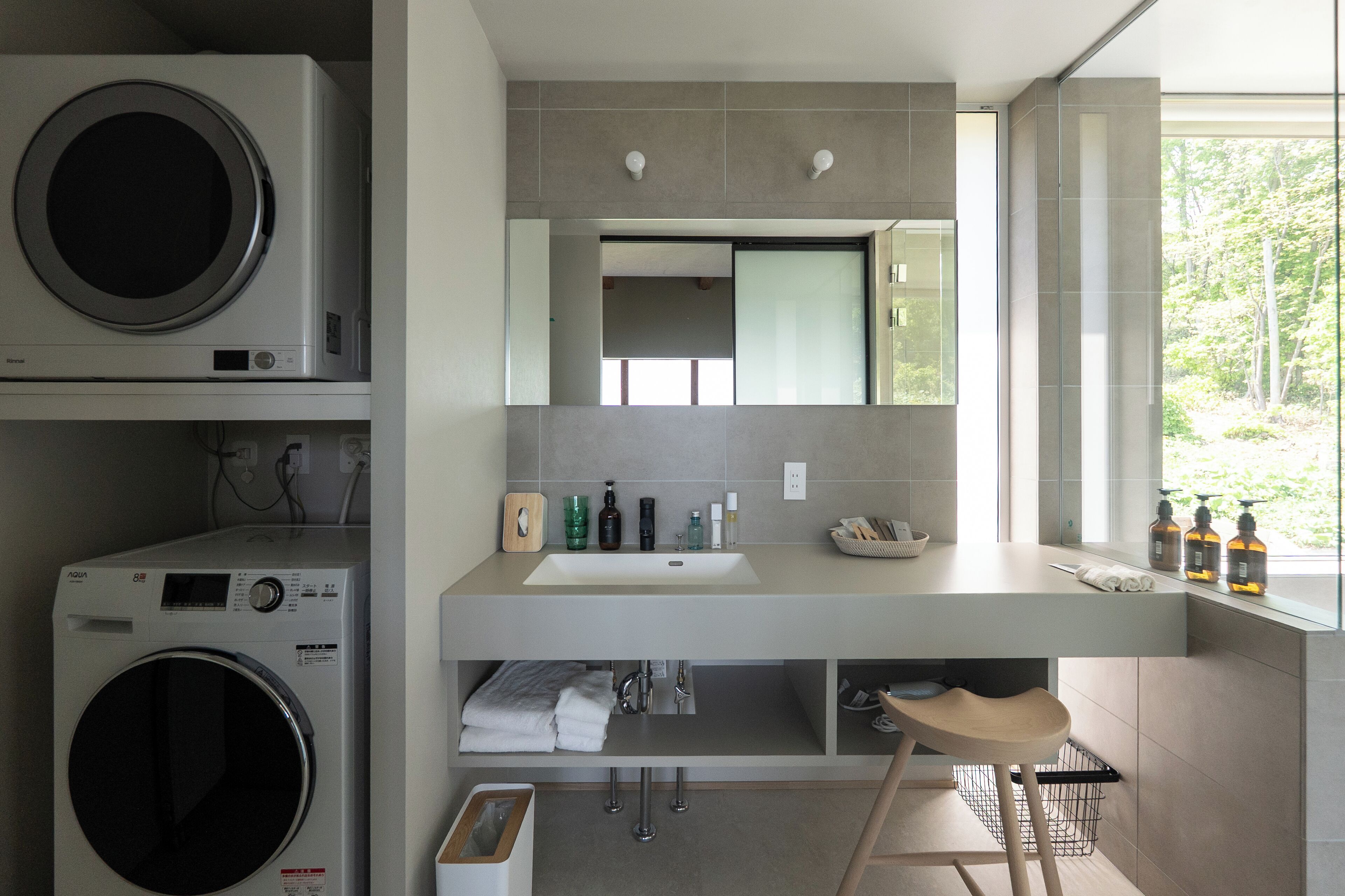 A modern bathroom with a built-in washing machine, a large mirror, and a window providing a view of trees.