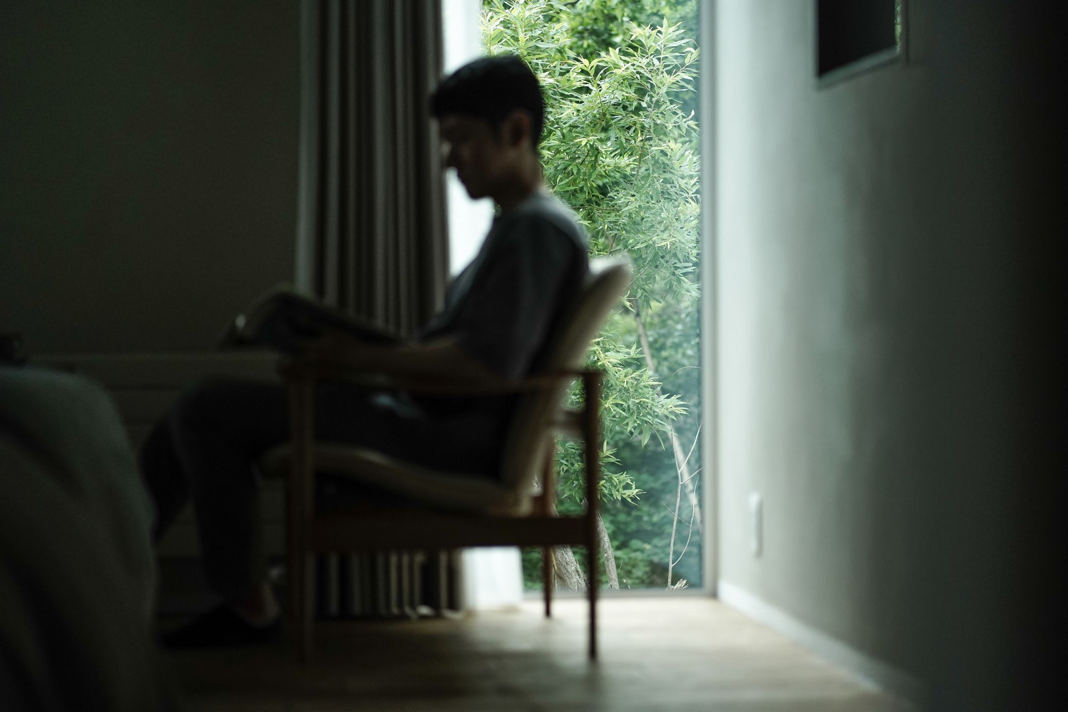 Eine Person sitzt auf einem Stuhl in einem Zimmer, durch das Fenster ist eine Pflanze zu sehen.