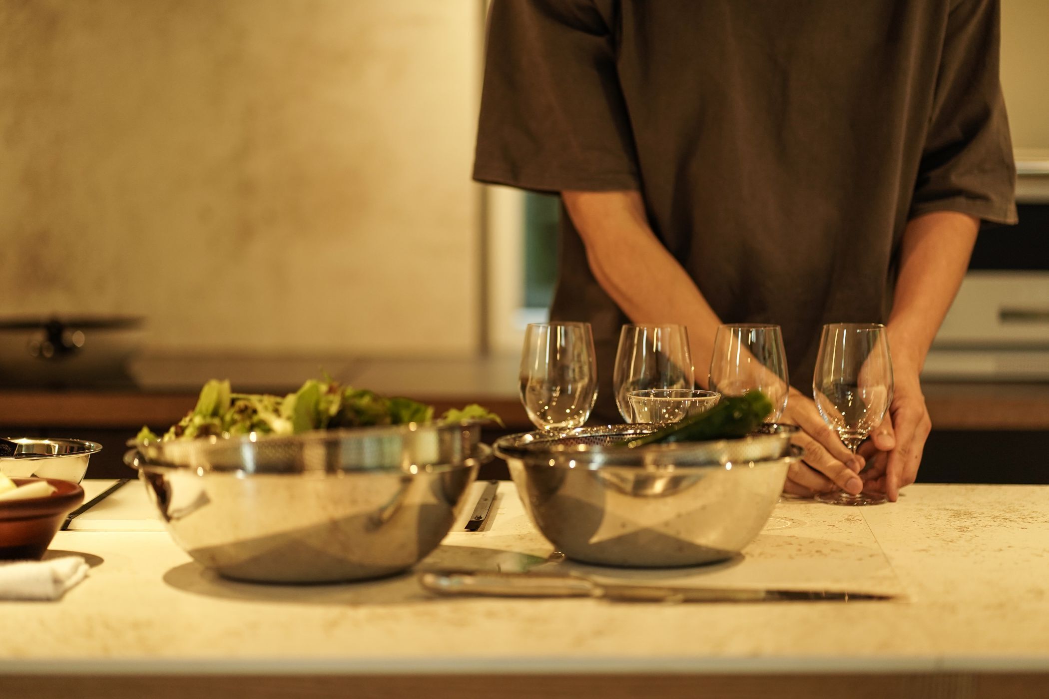 A kitchen counter with several stainless steel bowls containing food, a knife, and a person holding a wine glass.