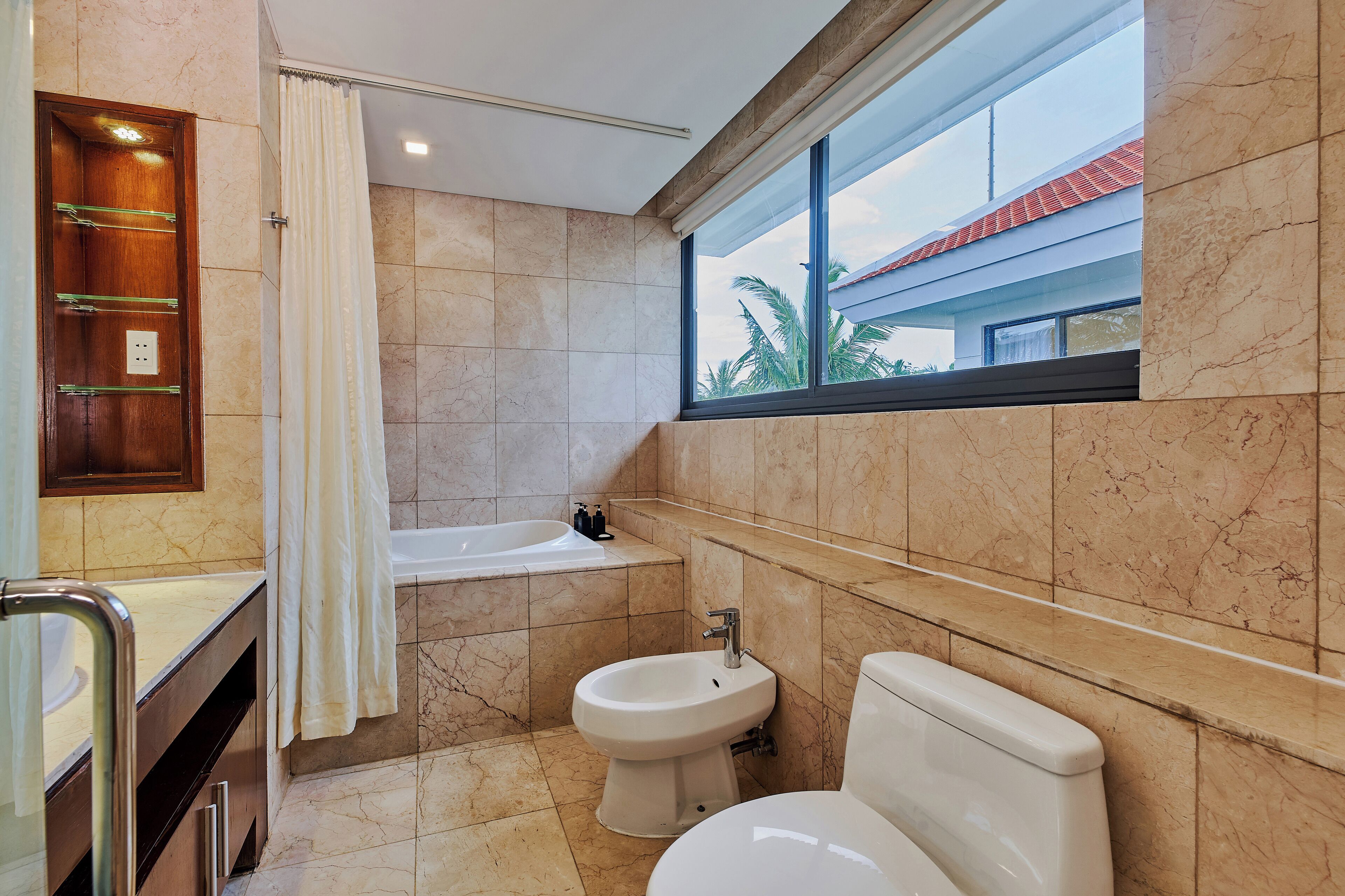 Un baño con bañera, bidé, inodoro y ventana con vista a palmeras.