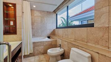Un baño con bañera, bidé, inodoro y ventana con vista a palmeras.