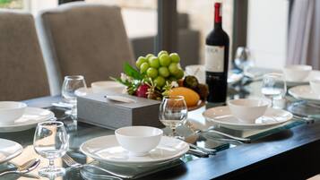 Une table à manger dressée avec de l’assiette, des bols et des couverts blancs, une bouteille de vin et une corbeille de fruits.