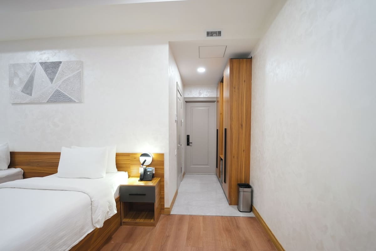A hotel room with a wooden bed, bedside table with a lamp, a small drawer, a trash bin, and a door leading to a hallway with wooden lockers.
