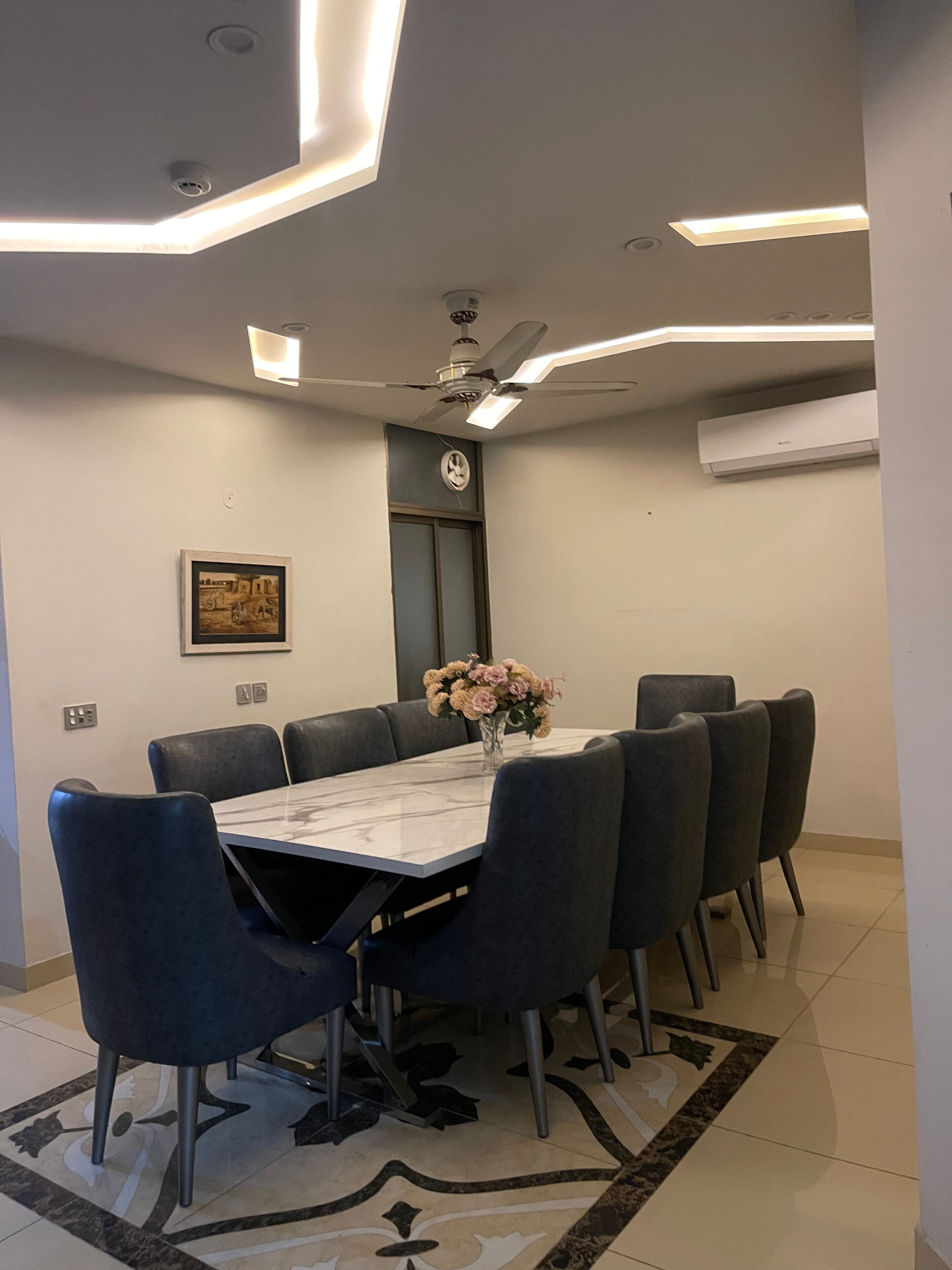 A modern dining area with a rectangular table, six chairs, a vase with flowers, a ceiling fan, recessed lighting, and an air conditioning unit.