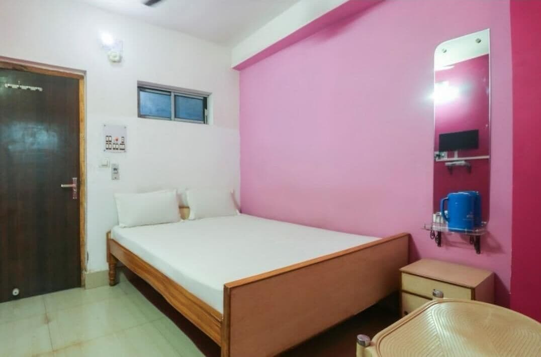 A bedroom with a single bed, a small bedside table, a chair, a window, a mirror, and a wall-mounted shelf with a blue container.