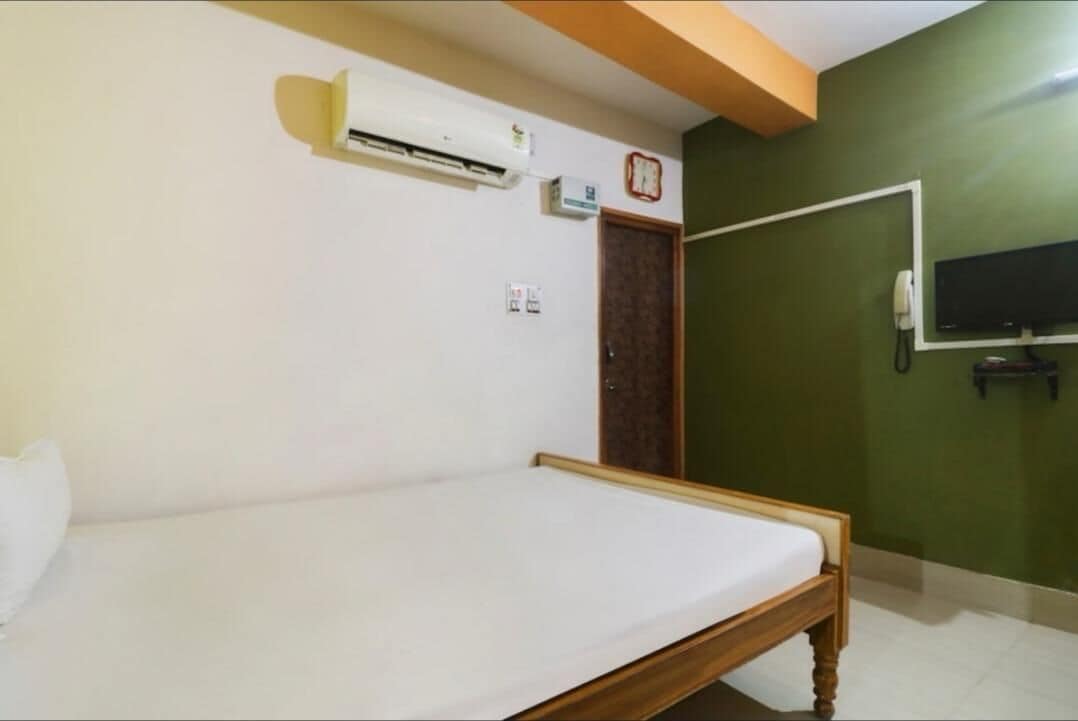 Una cama individual en una habitación con pared verde, un equipo de aire acondicionado, un reloj y un televisor montado en la pared.