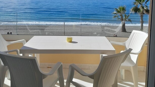Ein Balkon mit weißem Tisch und Stühlen, der auf einen Strand mit Palmen und das Meer blickt.