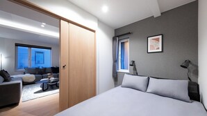 Ein modernes Schlafzimmer mit einem großen Bett, einem Nachttisch und einem Beistelltisch. Ein Fenster mit Vorhängen, ein gerahmtes Bild an der Wand und eine Tür zu einem anderen Raum sind ebenfalls zu sehen.