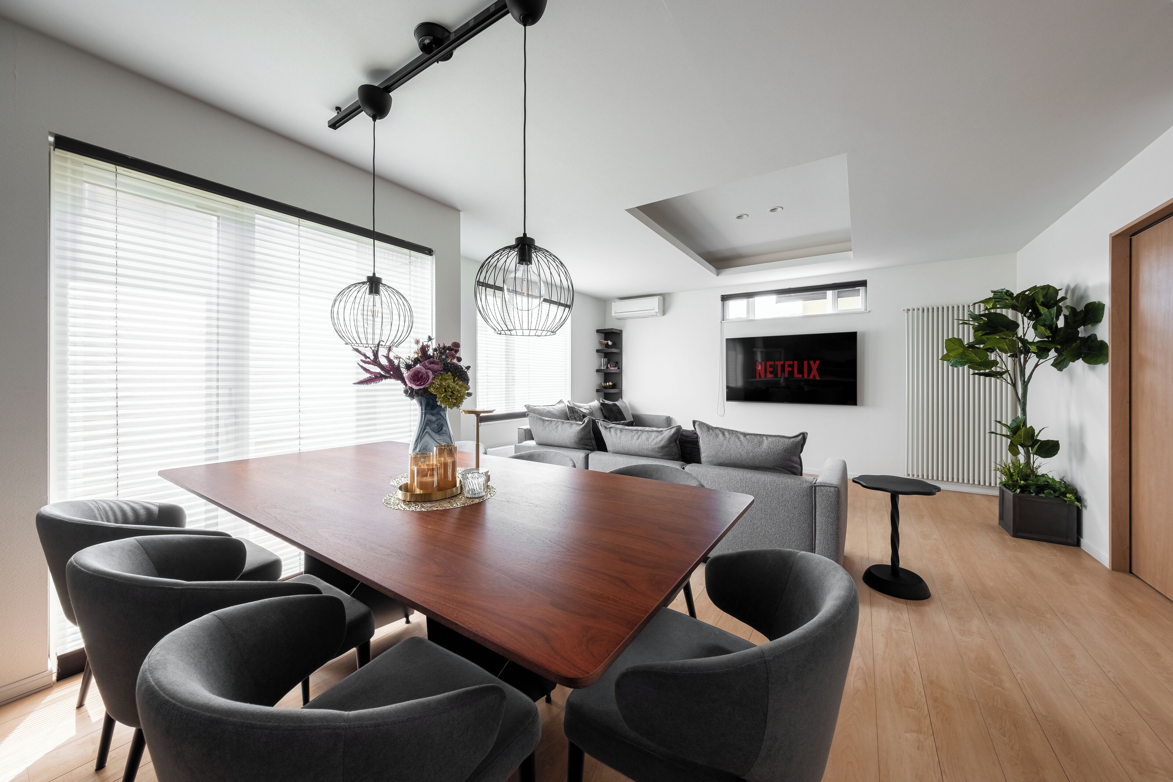 Una sala de estar moderna con una mesa de comedor de madera, sillas tapizadas en gris, un sofá, un televisor mostrando 'NETFLIX' y una planta en maceta.