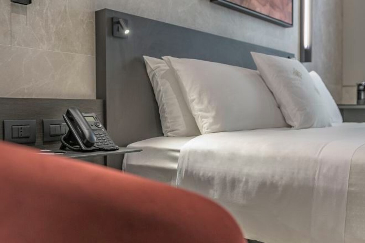 Una habitación de hotel con cama, almohadas blancas, un teléfono en la mesita de noche y una lámpara de pared.