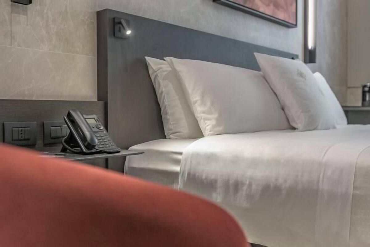 A hotel room with a bed, white pillows, a bedside telephone, and a wall-mounted light.