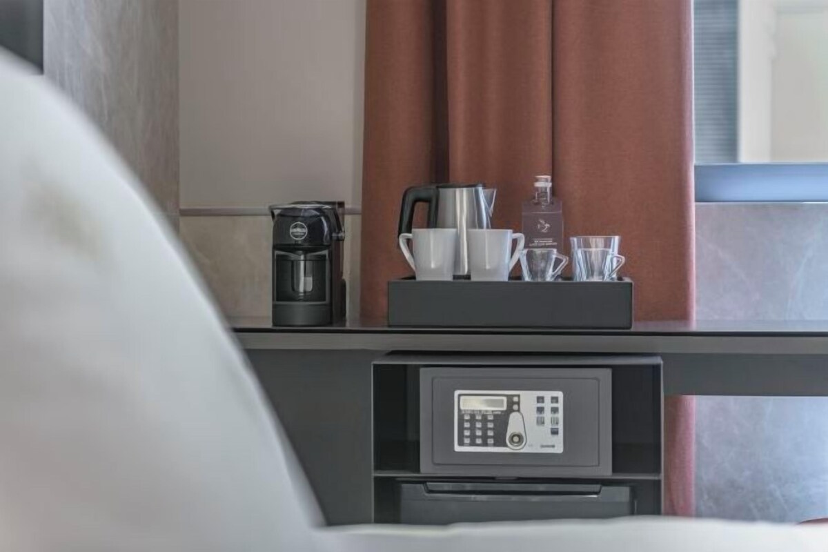 A hotel room with a coffee machine, a kettle, cups, and a glass set on a counter.