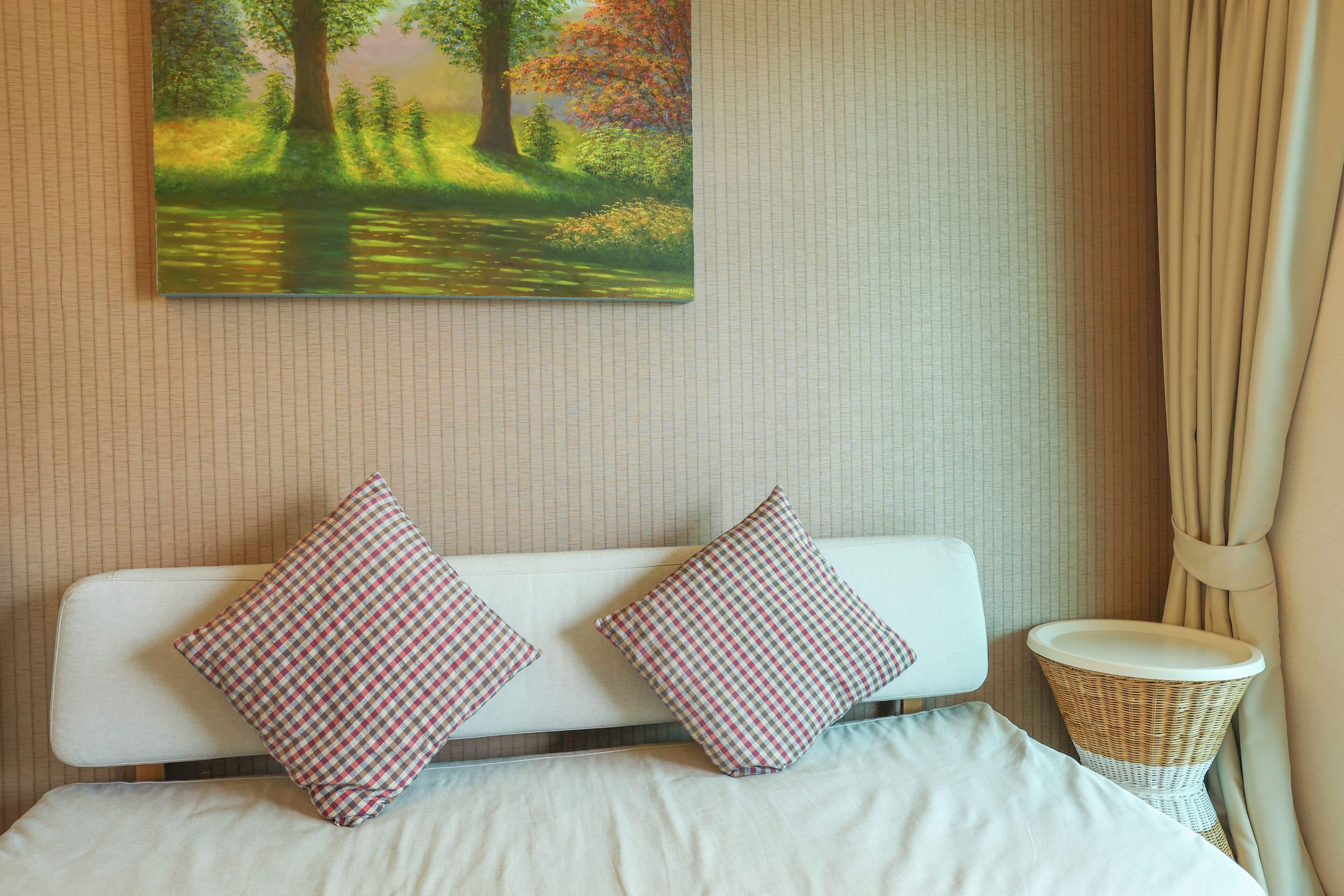 Una cama con dos almohadones a cuadros, una mesita de noche de mimbre y un cuadro de un paisaje forestal en la pared.