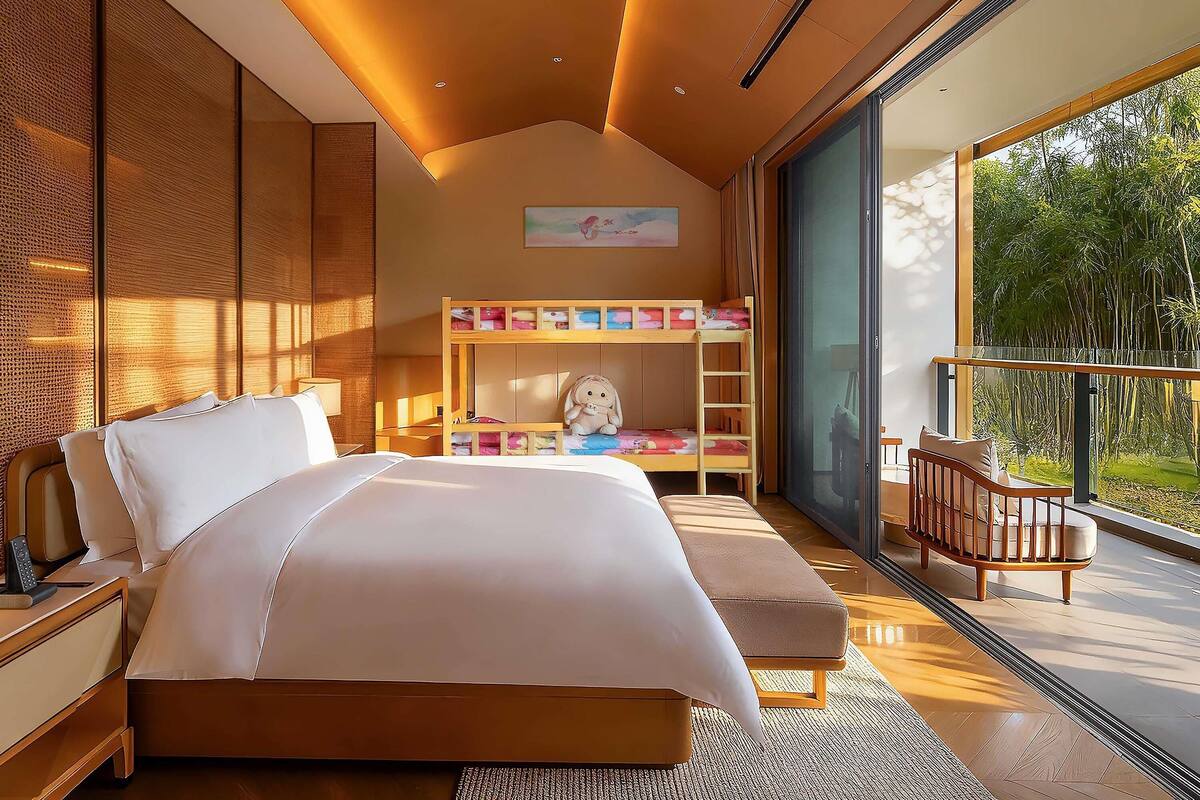 A modern bedroom with a large bed, a bunk bed, a balcony with a view of greenery, and a large window.