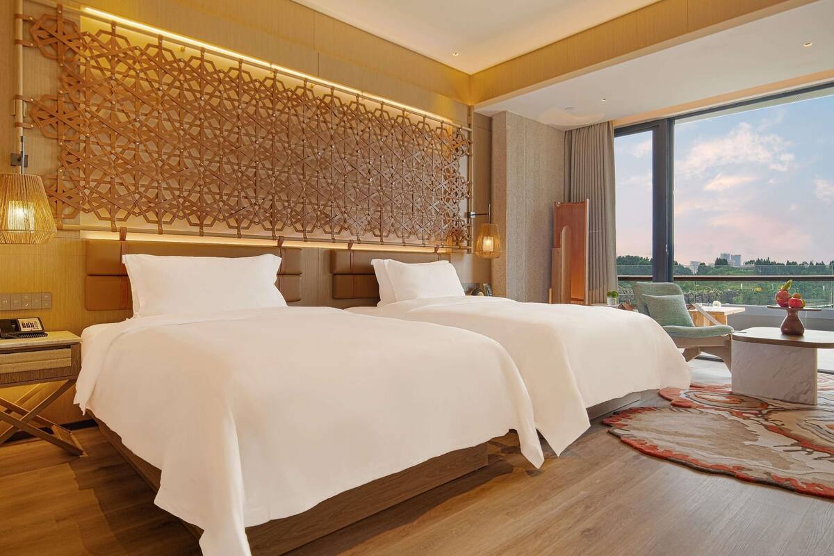 A hotel room with two beds, a large window, a decorative wall, and a small table with fruit.