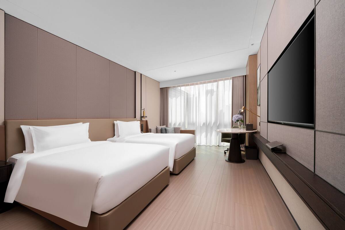 Une chambre d’hôtel avec deux lits, une télévision, un bureau et une grande fenêtre avec des rideaux.