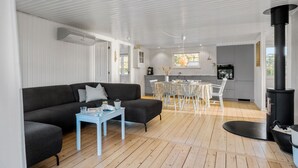 Ein modernes Wohnzimmer mit einem schwarzen Sofa, einem blauen Couchtisch und einem Essbereich mit weißem Tisch und Stühlen. In der Ecke ist ein schwarzer Kaminofen zu sehen.