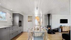 Eine moderne Küche mit weißen Schränken, einer Essecke mit rundem Tisch und blauen Stühlen, einem eingebauten Holzofen und einem an der Wand montierten Flachbildfernseher.
