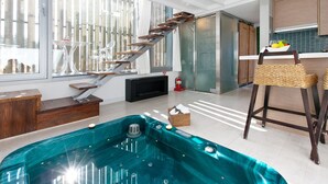 Una sala de estar moderna con una bañera de hidromasaje de fondo de cristal, una escalera de madera y una zona de cocina con una silla de mimbre.
