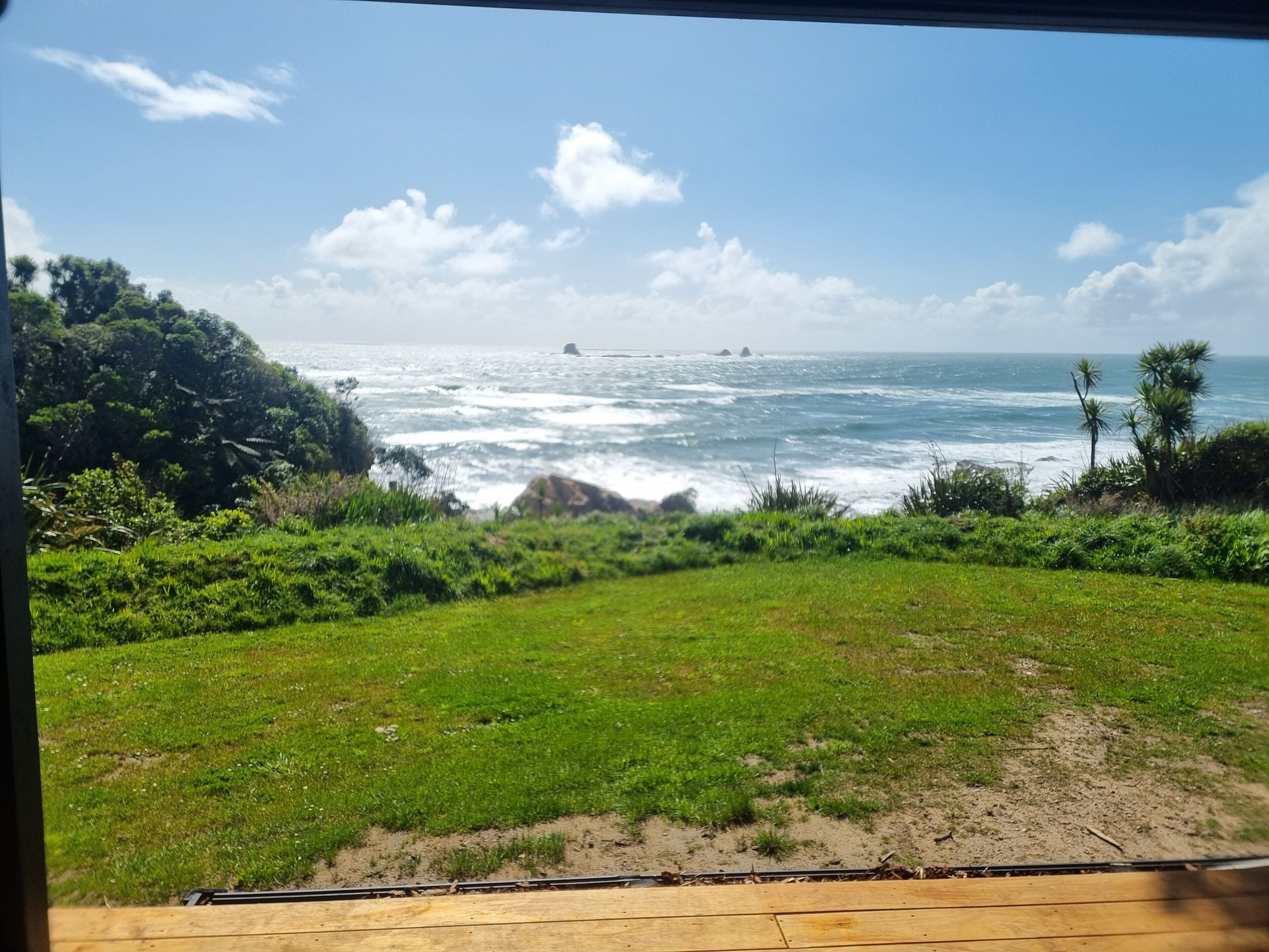 A coastal view with a grassy area, a wooden deck, and the ocean with waves and distant islands.