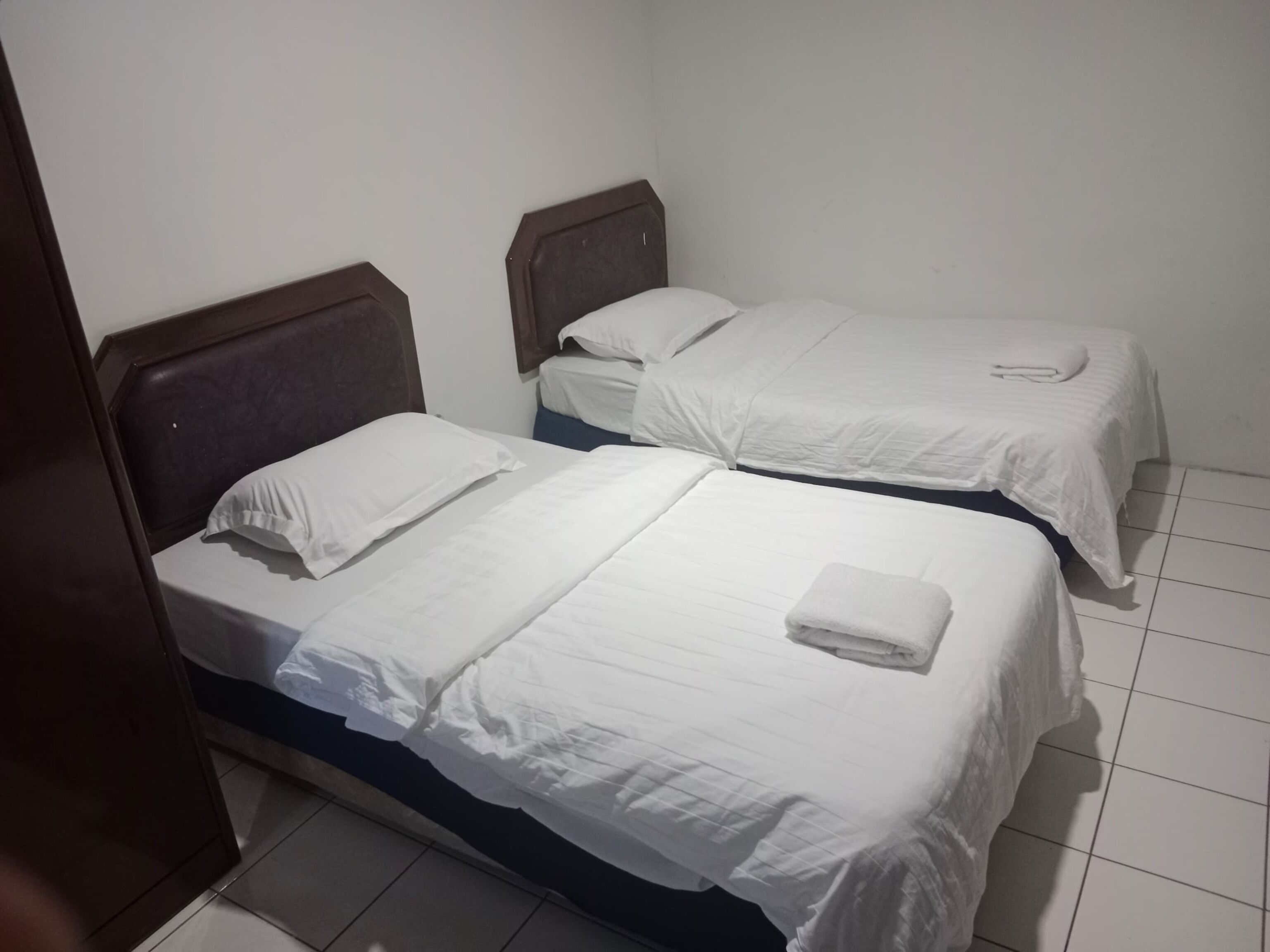 Dos camas individuales con cabeceros de madera en una habitación con paredes blancas y suelo de baldosas.