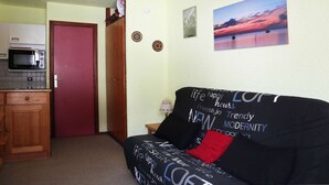 A compact living space with a kitchenette, a red door, a couch with motivational phrases, and a sunset painting. - Studio 4 pers. direct access slopes, center and south-facing balcony (Les Orres)