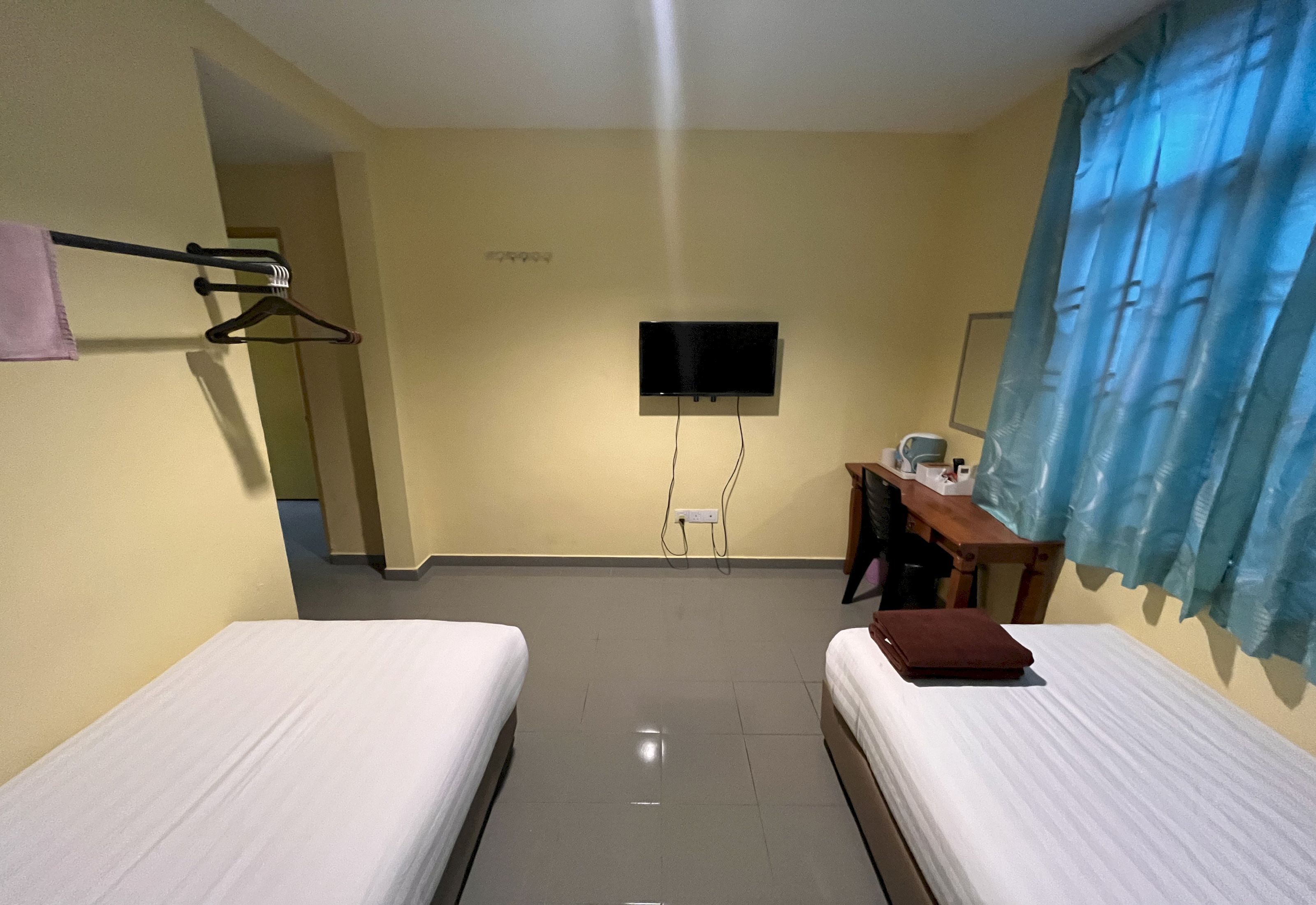 Ein Hotelzimmer mit zwei Betten, einem an der Wand befestigten Fernseher, einem Schreibtisch mit Stuhl und einem Fenster mit blauen Vorhängen.