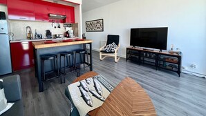 A modern kitchen with red cabinets, a central island with stools, a ceiling fan, a dining area with a TV, and a patterned throw on the sofa.