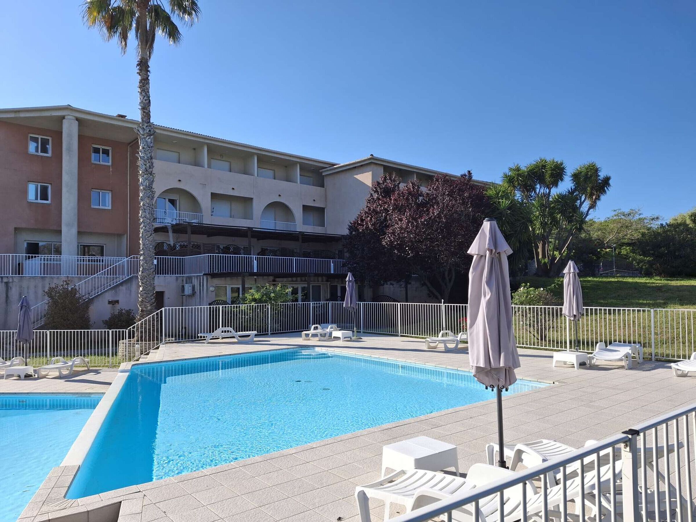 Una piscina con sillas de descanso y sombrillas, rodeada por una cerca blanca, con un edificio al fondo.