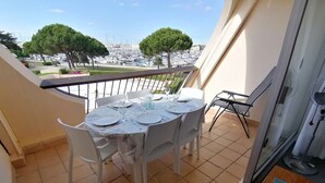 A balcony with a round table set for dining, overlooking a marina with boats and trees. - Renovated 2-room apartment with pool and parking (Port Camargue)