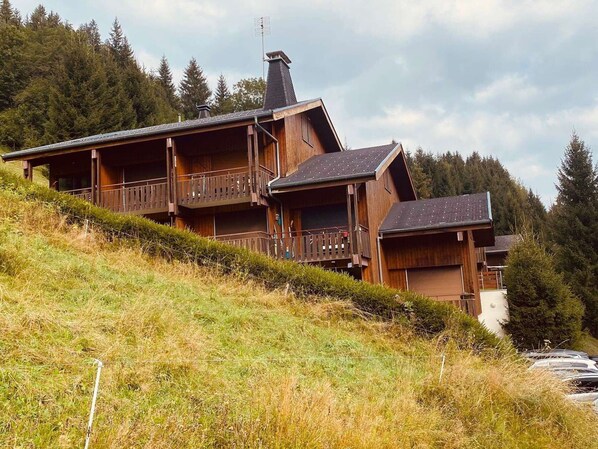 Una cabaña de madera con varios balcones, ubicada en una ladera con un bosque de fondo.