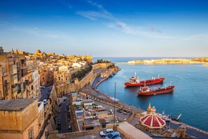 A coastal cityscape with historic buildings, a fortified wall, and two large red ships docked at the harbor. - Kenn Gozitan Villa and Pool (Gharb)