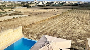 A view of a swimming pool area with a beige umbrella, surrounded by dry fields and a distant townscape.