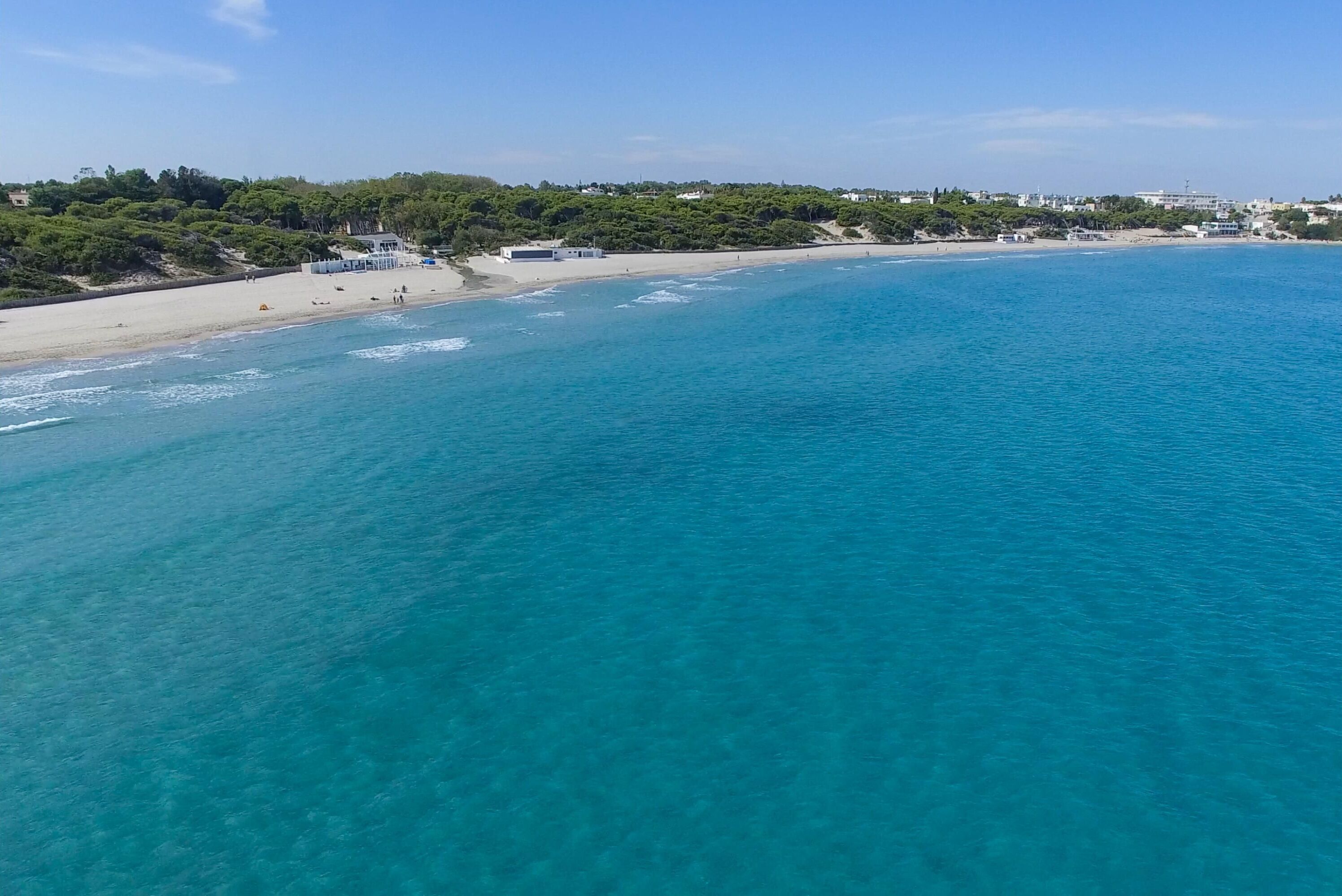 A coastal beach with clear turquoise waters, a sandy shore, and buildings in the background.