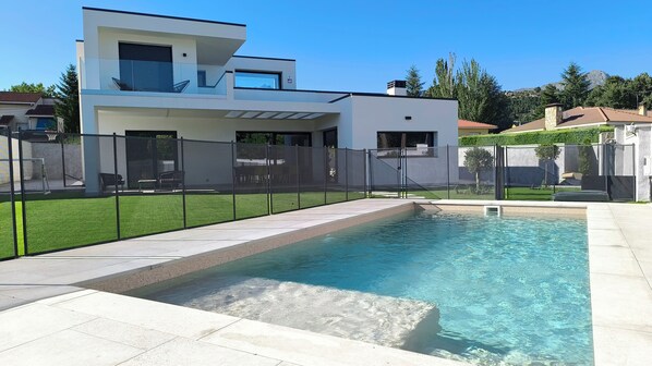 Una casa moderna con piscina, patio trasero cercado y cielo azul despejado.