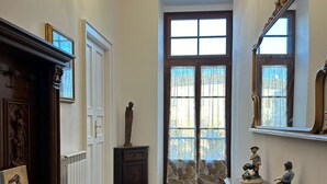 A hallway with a chandelier, wooden furniture, a large window with curtains, and a statue. - Camera famigliare un letto matrimoniale e 1 lettini (Corropoli)