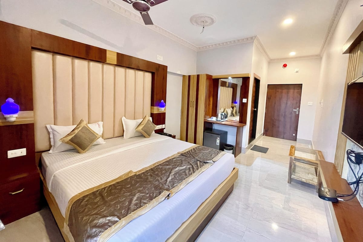 A hotel room with a large bed, a ceiling fan, a desk with a mirror, and a small kitchenette.