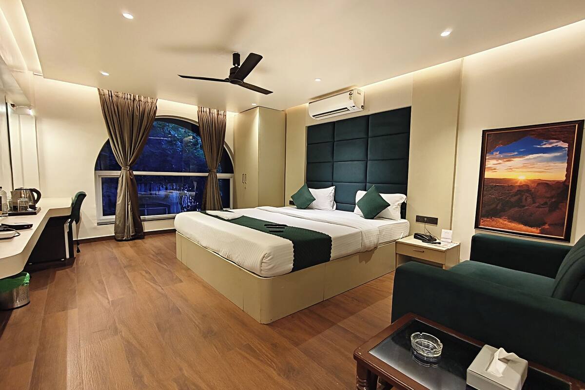 Quarto de hotel moderno com uma cama grande, uma escrivaninha, uma cadeira, um sofá, um ventilador de teto, um ar condicionado e uma janela grande com cortinas.