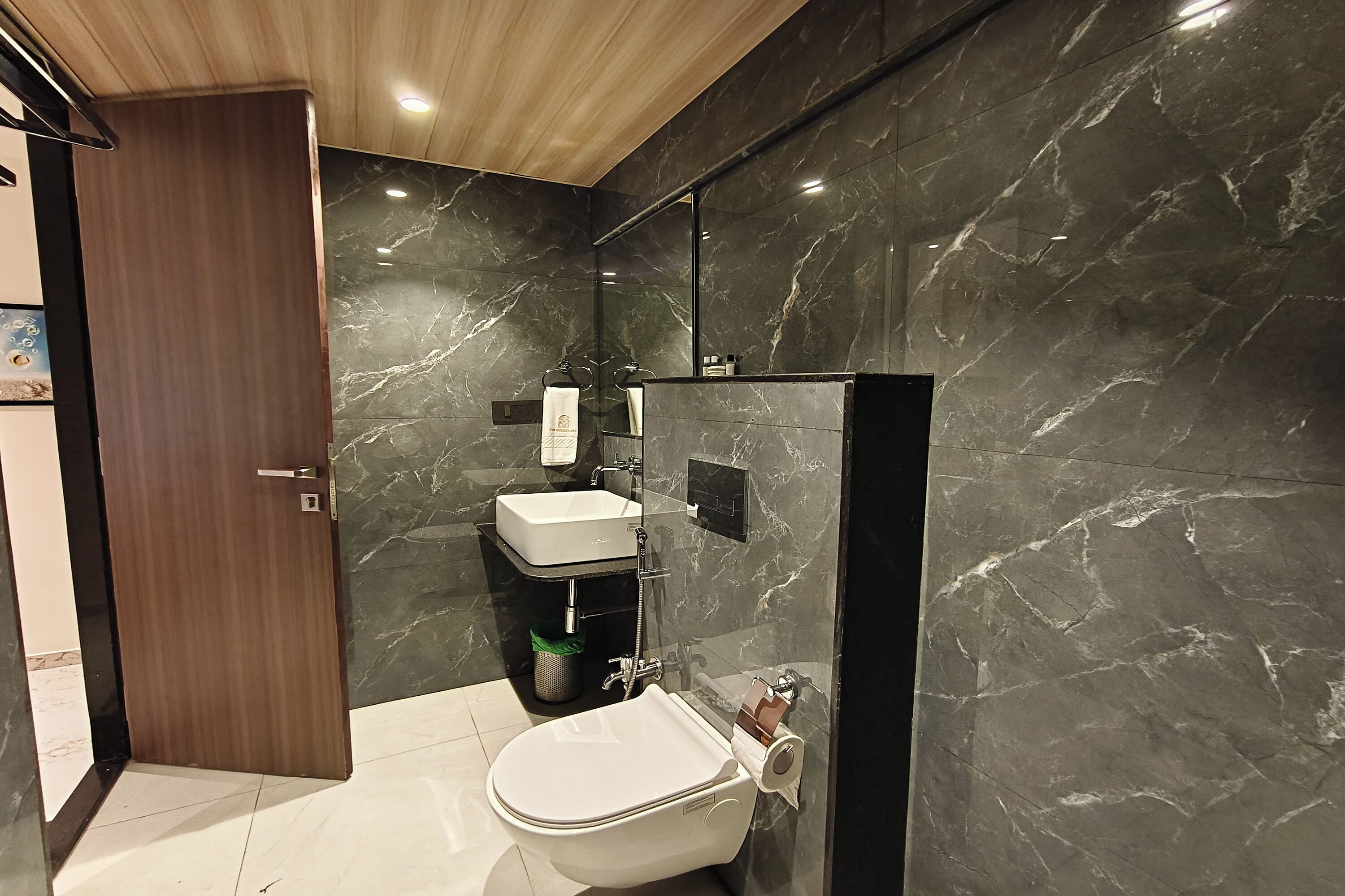 A modern bathroom with a marble wall, a wooden door, a white sink, and a toilet.