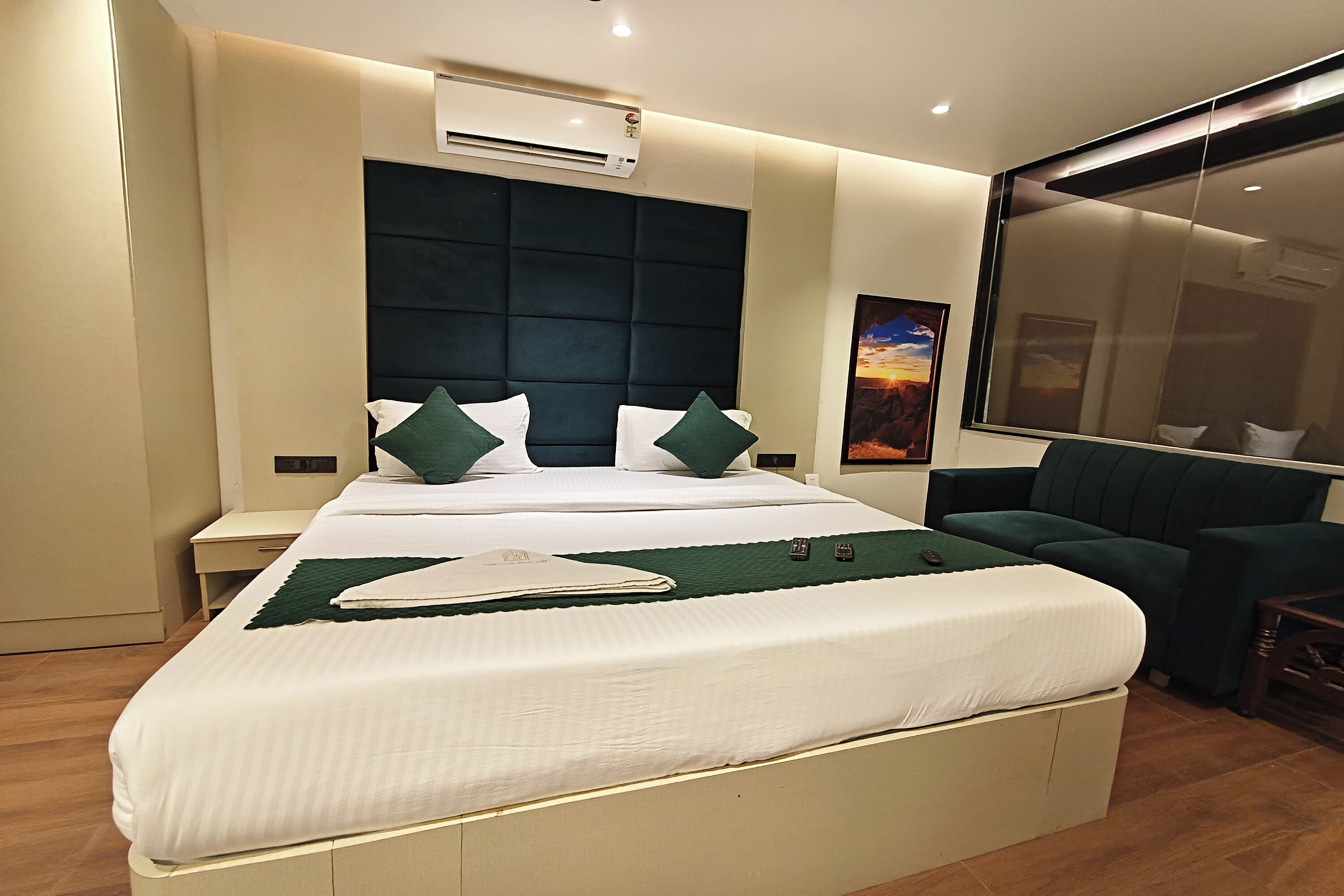 A modern hotel room with a large bed, a dark headboard, a bedside table, a small desk, a green sofa, and a large window.